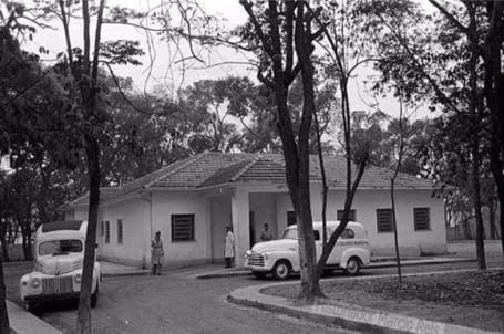 A foto preto-e-branco mostra uma casa cercada de árvores; em frente a ela estão duas ambulâncias de modelo antigo, dos anos 50